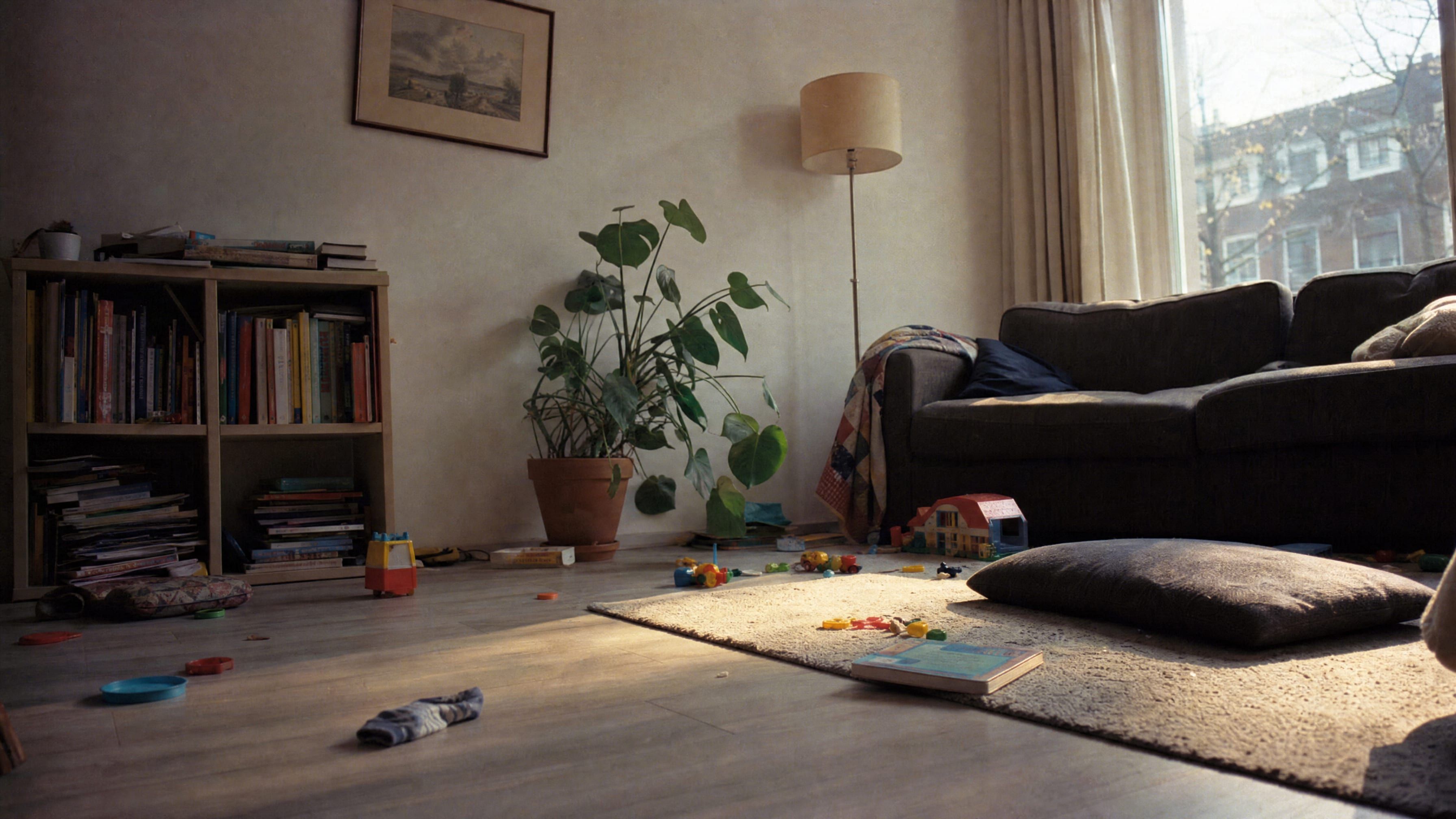 Een woonkamerhoek in een gewone woning, laag bij de vloer gezien: een laminaatvloer met speelgoed erover verspreid, een kleedje in laag zonlicht, een bank met een plaid en een boekenkast tegen de wand.